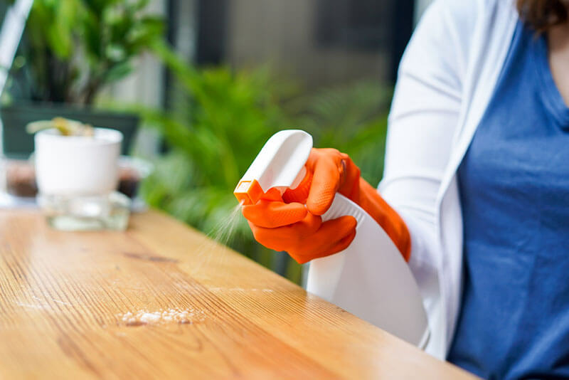 woman wearing orange gloves is cleaning a table using a spray bottle